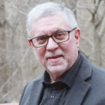 An older man with gray hair, glasses, and a beard wearing a suit jacket, standing outdoors in winter.