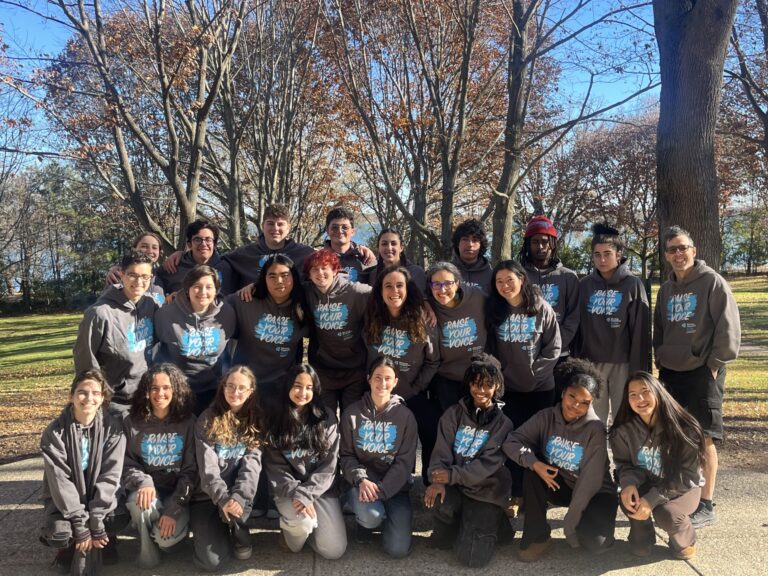A group of young adults in matching hoodies pose together outside on a sunny autumn day with bare trees behind them.