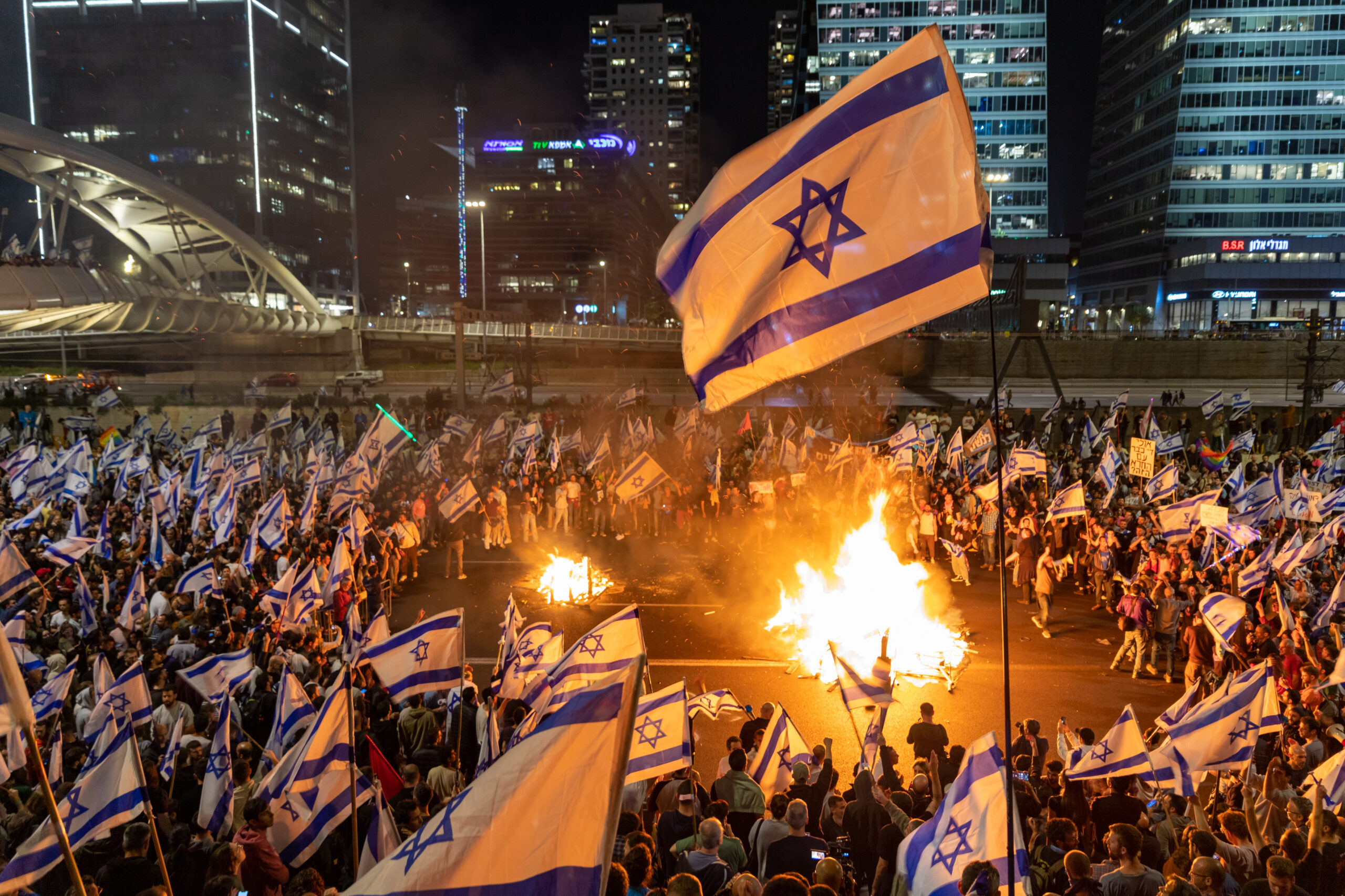 Large crowd waving Israeli flags gathers at night by burning fires in a city street surrounded by tall buildings.