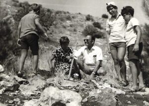 Four boys and a man outdoors on rocky terrain, smiling and talking on a sunny day.