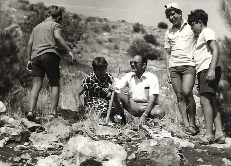 Four boys and a man outdoors on rocky terrain, smiling and talking on a sunny day.