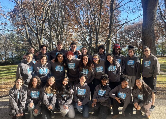 A group of young adults in matching hoodies pose together outside on a sunny autumn day with bare trees behind them.