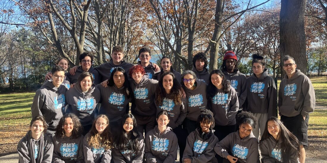 A group of young adults in matching hoodies pose together outside on a sunny autumn day with bare trees behind them.