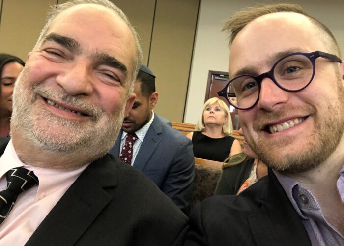 Two smiling individuals in suits take a selfie at an indoor event, with people seated in the background.