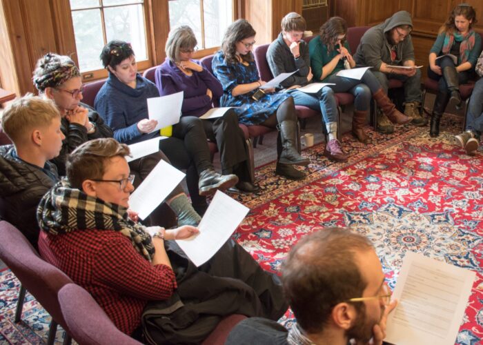 A group of people sit in a circle on chairs, reading papers in a cozy, wood-paneled room with large windows.