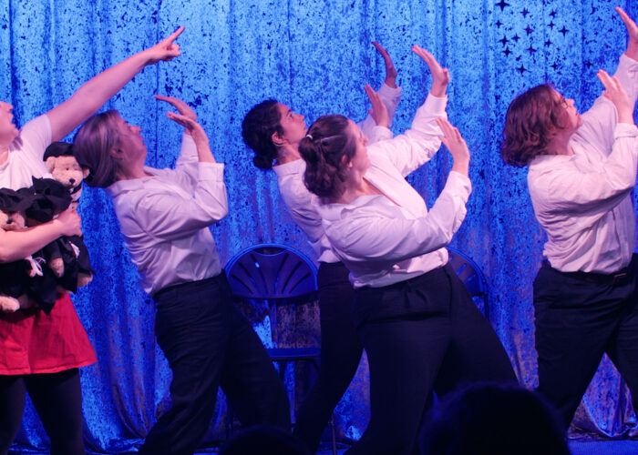 Five performers in white shirts pose dramatically under blue stage lights. One holds a stuffed animal.