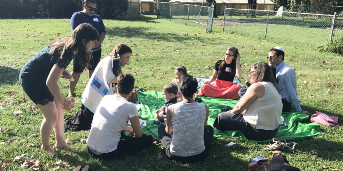 A group of people sit on a blanket outdoors in the sun, listening to a woman holding a colorful chart.