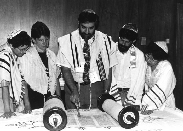 Group studying a Torah scroll on a table, with a man pointing at the text.