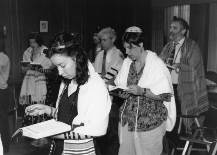 People wearing prayer shawls and head coverings, reading from books in a dimly lit room.
