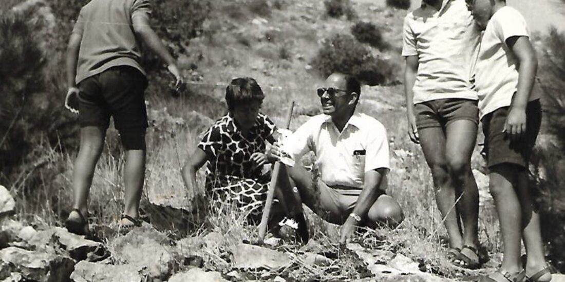 Four boys and a man outdoors on rocky terrain, smiling and talking on a sunny day.