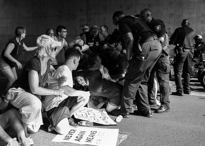 Protesters sitting on the ground as police officers detain someone; protest signs are visible.