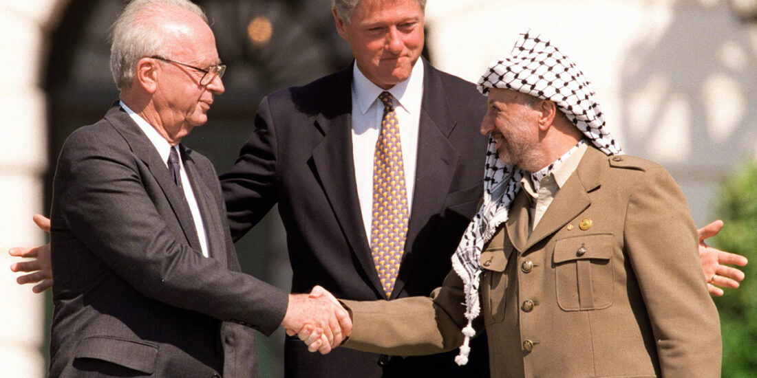 Three men in suits and uniform shake hands outdoors, with one man standing between them, smiling and guiding the handshake.