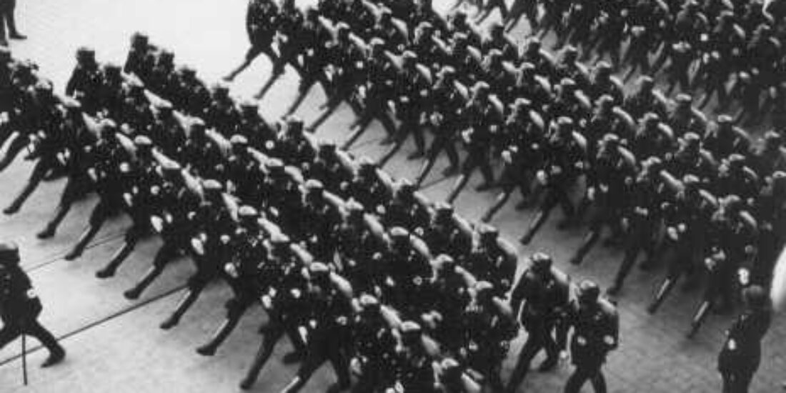 A group of uniformed soldiers march in formation on a paved street, viewed from above.