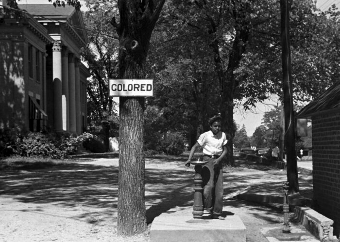 A Black boy stands at a water fountain beneath a sign reading COLORED in a segregated area.