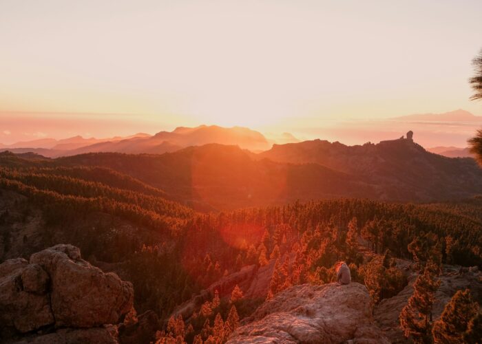 Sunset over a forested mountain landscape with a person sitting on rocks in the foreground.