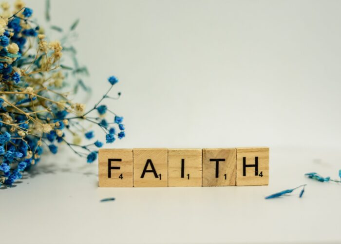 Scrabble tiles spell FAITH beside a bunch of dried blue and beige flowers on a white background.