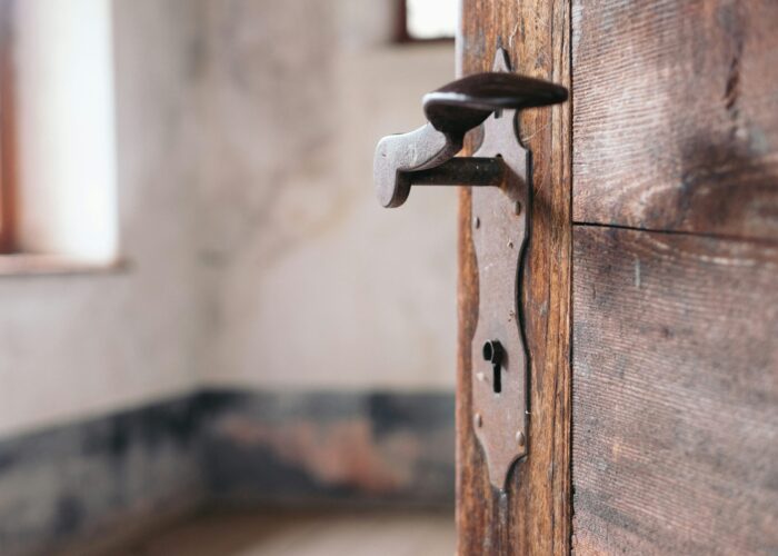 Close-up of an old wooden door with a vintage handle and keyhole, leading into an empty, blurred room.