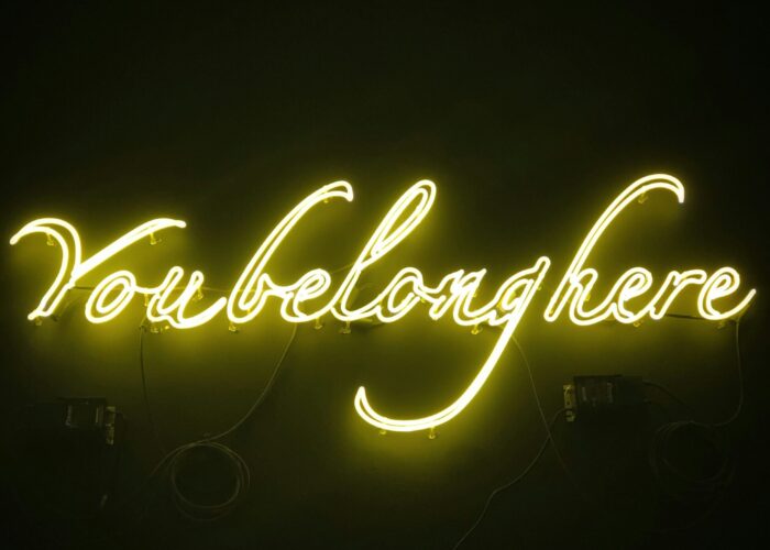 Neon sign in cursive script that reads You belong here glowing brightly against a dark background.