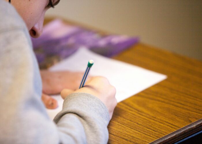 A person in a grey sweatshirt writes with a pencil on paper at a wooden desk.