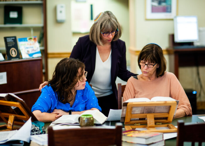 Three women sitting at a table in a library, reading books and discussing.