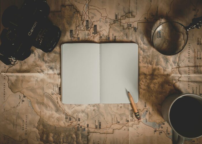 Open notebook, pencil, camera, magnifying glass, and coffee on a vintage map, viewed from above.