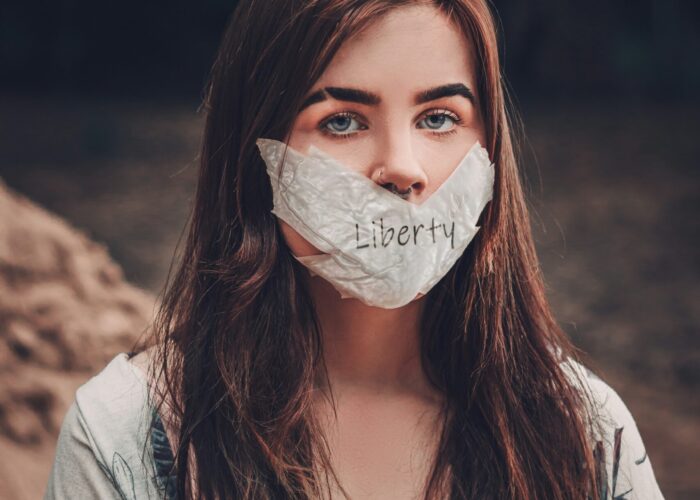 Woman with long brown hair wears a cloth labeled Liberty covering her mouth, sitting outdoors.