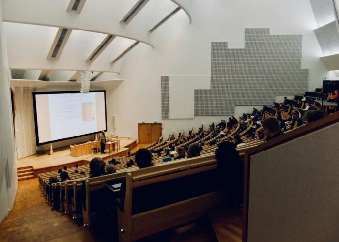 A large lecture hall with students seated and a presentation projected at the front.