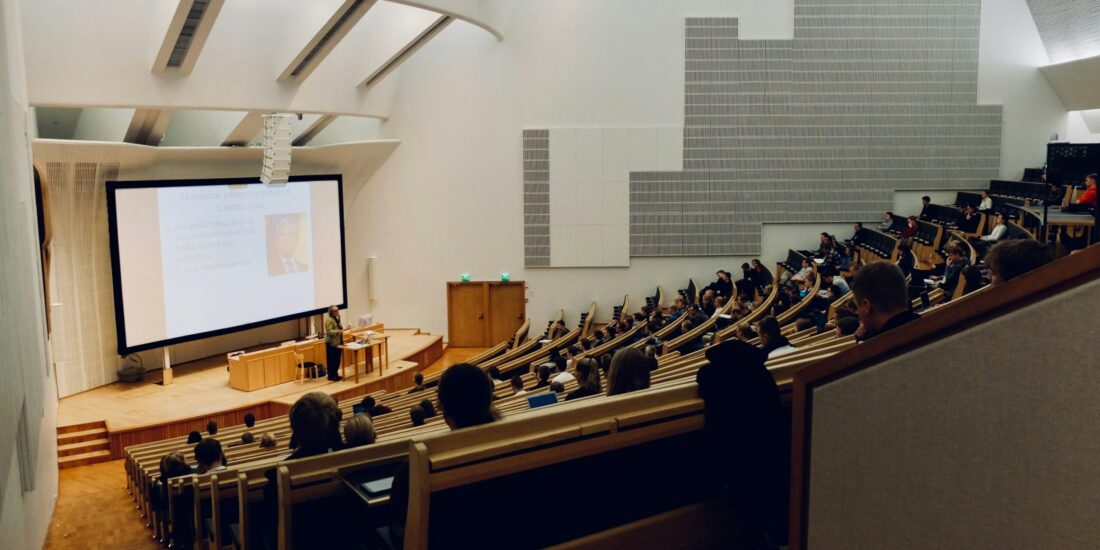 A large lecture hall with students seated and a presentation projected at the front.