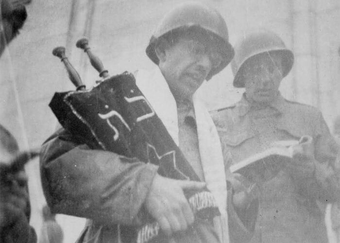 A soldier in a helmet holding a Torah scroll, accompanied by another soldier in uniform, both are reading.
