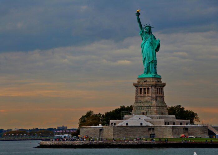 The Statue of Liberty stands on its pedestal with a cloudy sky in the background and water in the foreground.