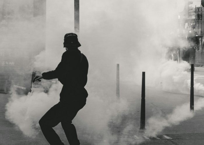 Person in hat and jacket runs through thick smoke on a city street.