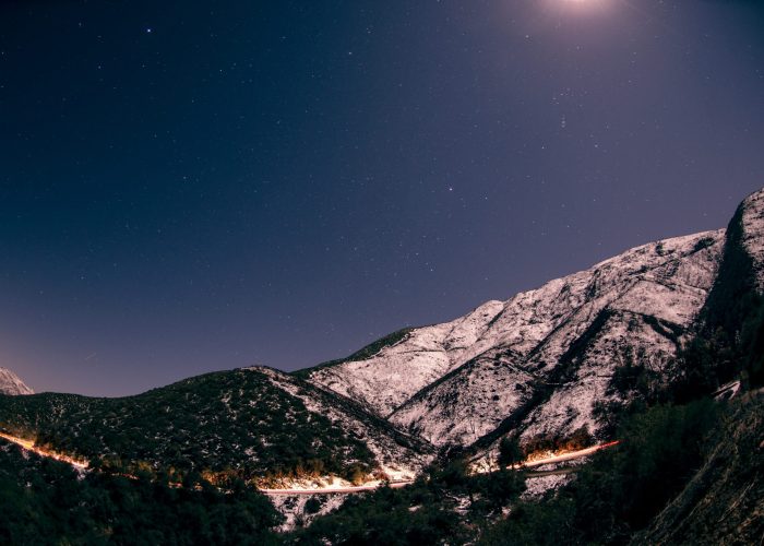 Starry night sky over snow-covered mountains with a faint light trail from a distant road below.