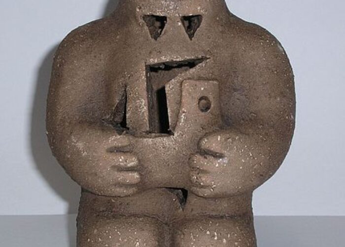 Clay sculpture of a golem with a triangular head holding an object, against a light gray background.