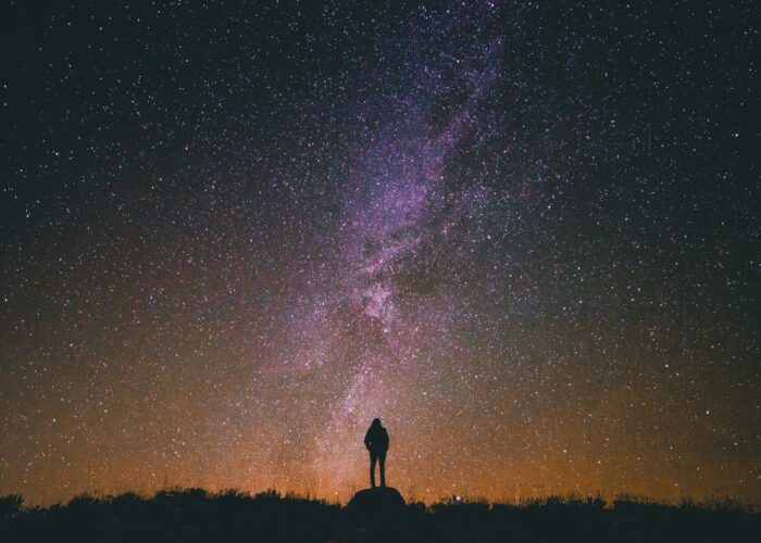 A person stands on a hill silhouetted against a star-filled night sky and the Milky Way.