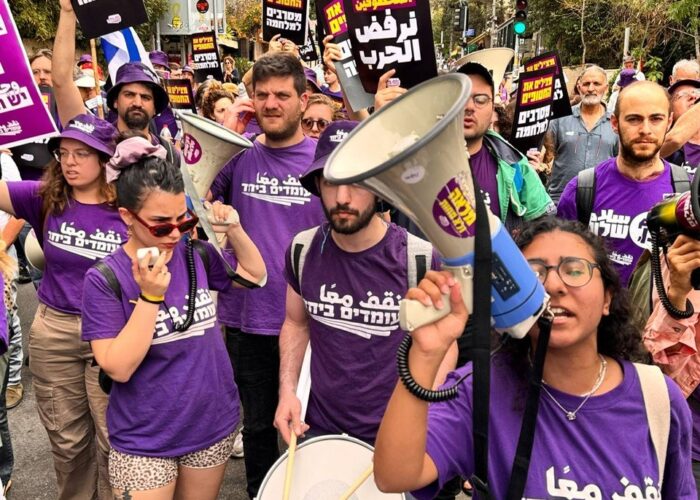 Protesters wearing purple shirts hold signs and megaphones, participating in a street demonstration.