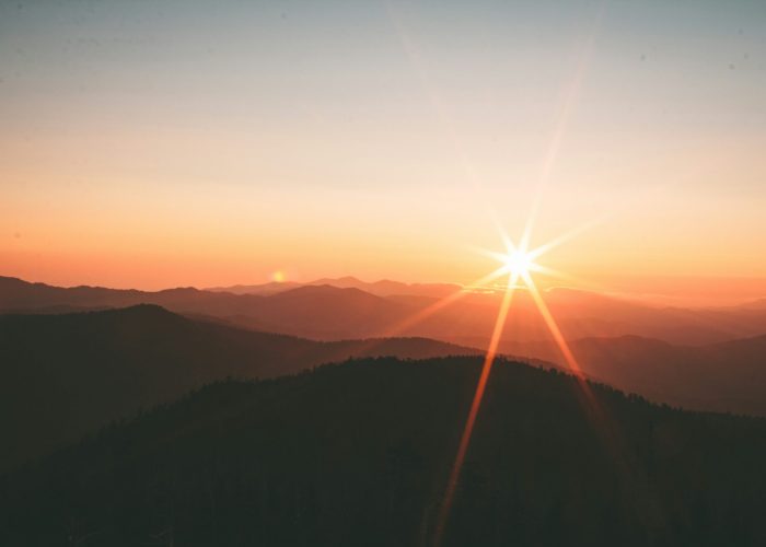 Sunrise over mountains with a bright sunburst, soft orange sky, and layered silhouettes of hills.