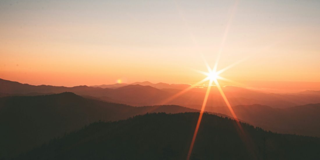 Sunrise over mountains with a bright sunburst, soft orange sky, and layered silhouettes of hills.