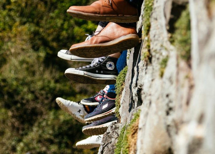 Six pairs of legs in different shoes dangle over a stone wall, with greenery and a house roof in the background.