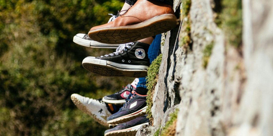 Six pairs of legs in different shoes dangle over a stone wall, with greenery and a house roof in the background.