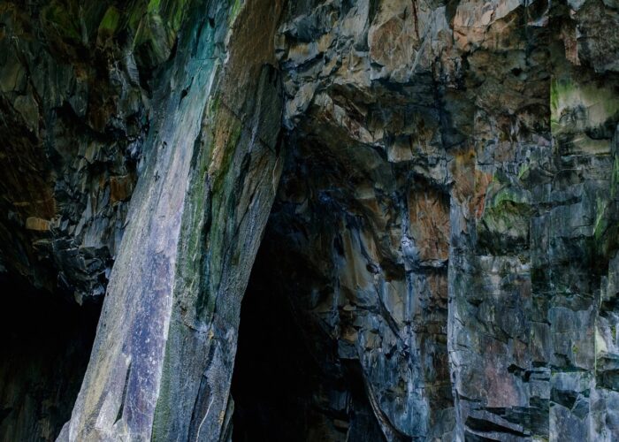 Natural cave entrance with rocky walls and a shallow pool of water at the base, surrounded by textured stone surfaces.