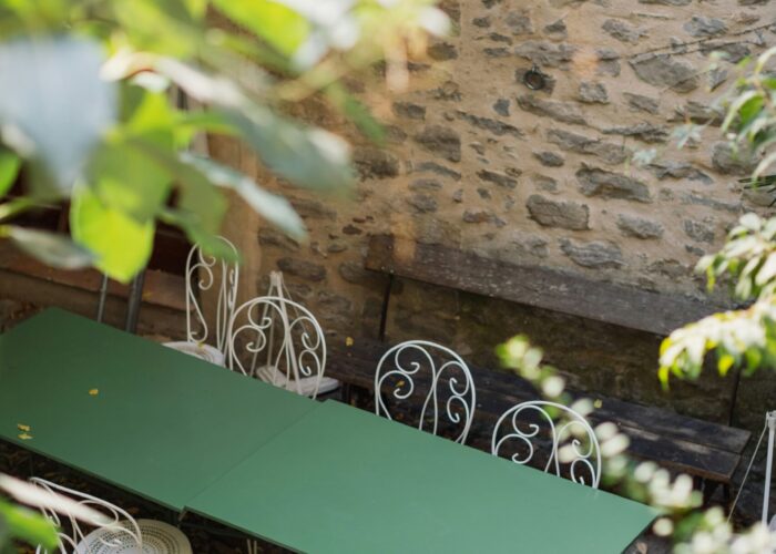 A green table with white chairs in a cozy outdoor courtyard surrounded by greenery and stone walls.