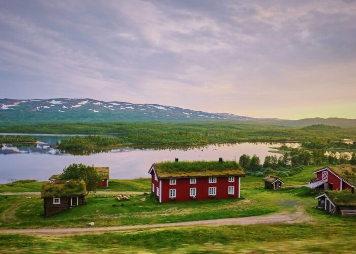 Red houses with grassy roofs beside a lake and mountains under a cloudy sky.