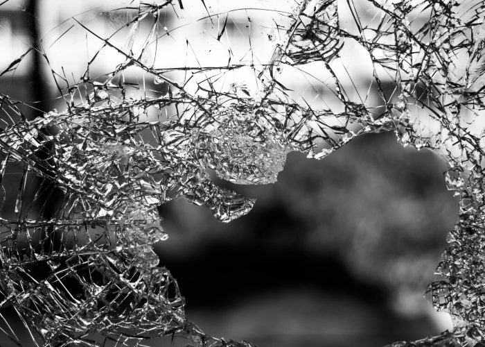 Close-up of shattered glass with jagged edges and a large hole, set against a blurred outdoor background.