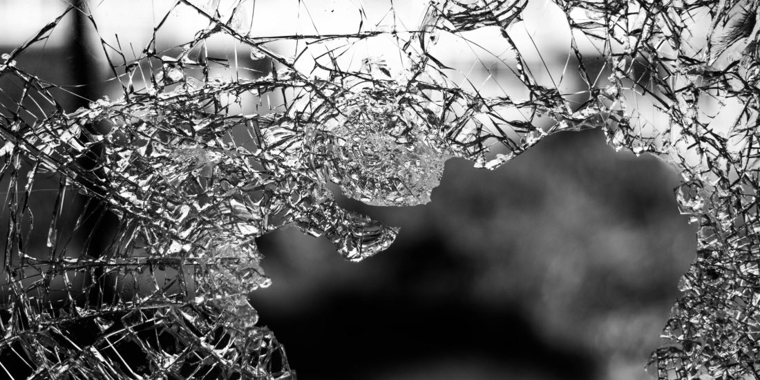 Close-up of shattered glass with jagged edges and a large hole, set against a blurred outdoor background.