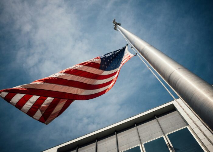 View from below of a U.S. flag waving on a tall flagpole against a blue sky, with a building partially visible.