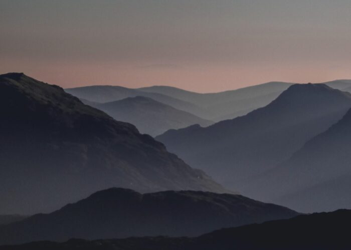 A serene view of layered mountain silhouettes at dusk, with a gradient sky transitioning from dark to light.