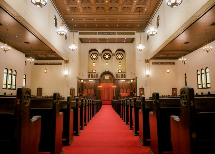 Interior of a synagogue with wooden pews, red carpet aisle, and ornate ceiling with hanging lights.