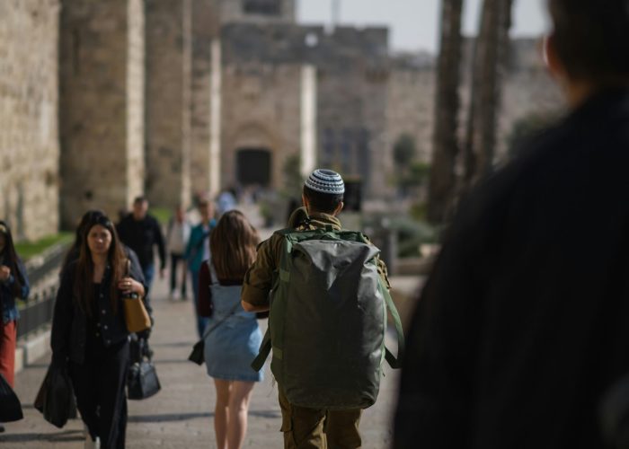 A soldier with a large backpack walks along a stone path, surrounded by people and palm trees.