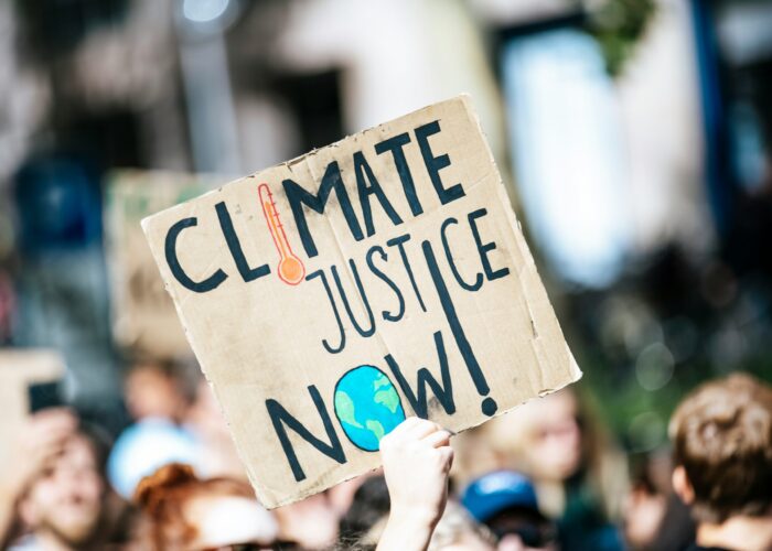 Protest sign reads CLIMATE JUSTICE NOW! with a thermometer and Earth illustration in a crowd.