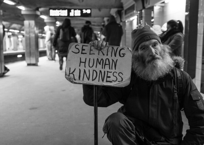 Man in a subway station holds a sign reading Seeking Human Kindness; people walk in the background.